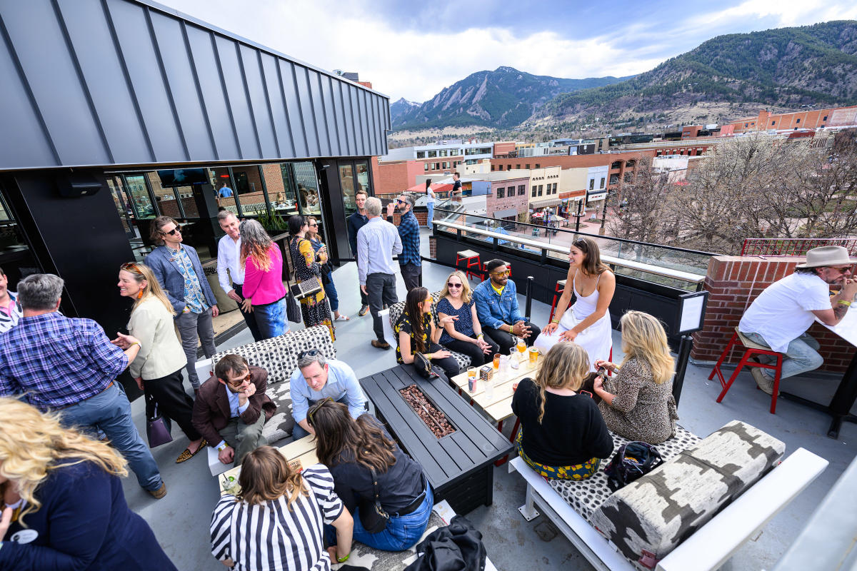 A group of businesspeople gather in small groups on a rooftop with views of the Flatirons.