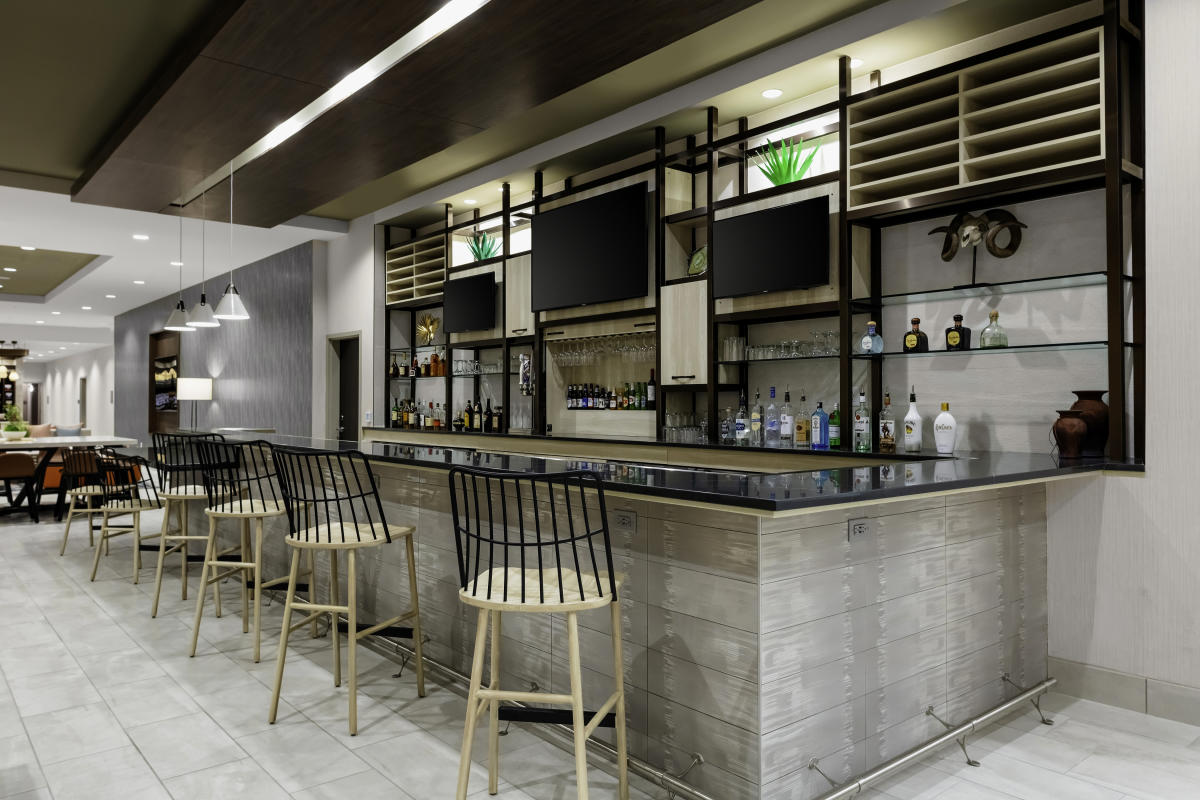 Modern style bar with black metal shelving holding various alcohol bottles, modern wood bar stools pushed against the L-shaped bar with black counter tops, and 3 TVs mounted in between bar shelving on the wall.