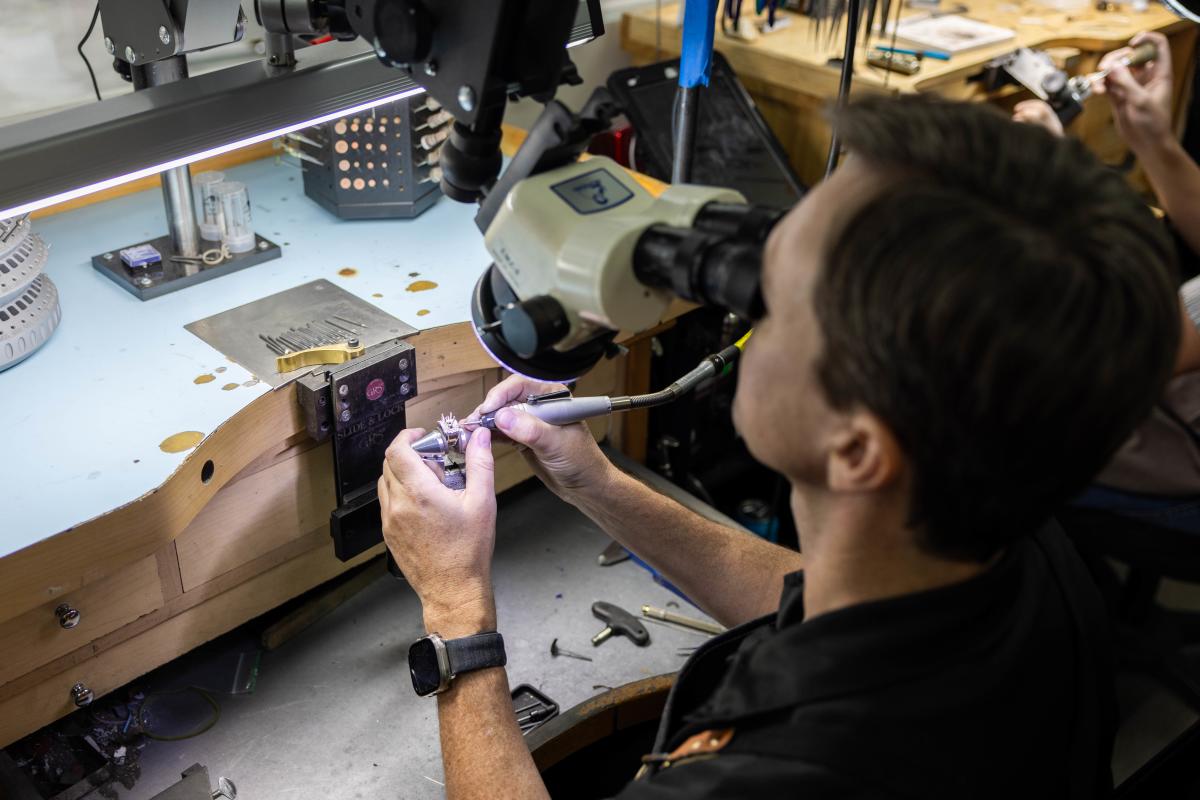 An over-the-shoulder view of a master goldsmith setting diamonds in an engagement ring at his desk at Christopher Fine Diamonds in Chandler, AZ, using jeweler's tools.