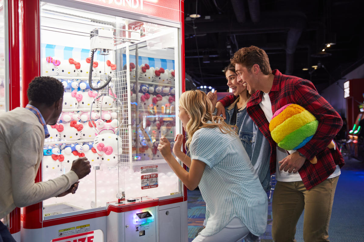 Group of 4 adults standing crowding around a claw machine with excitement as the claw is beginning to pick up a light pink Hello Kitty plushy.