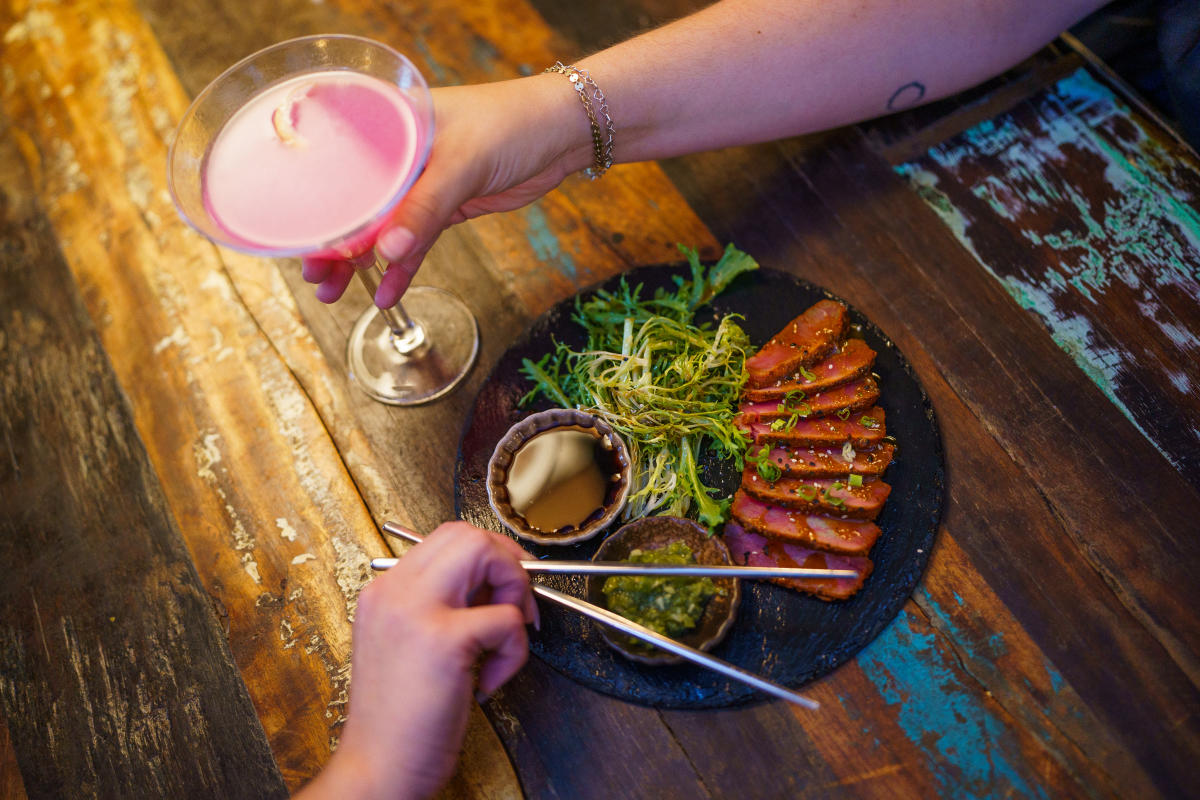 A hand holding a pink martini out on a wooden table with another hand holding chopsticks above an appetizer.
