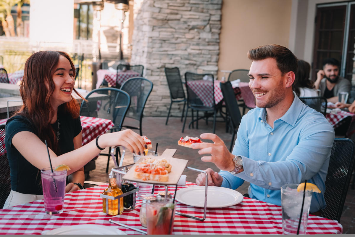 Couple sitting on patio at table picking up appetizer to eat and smiling.
