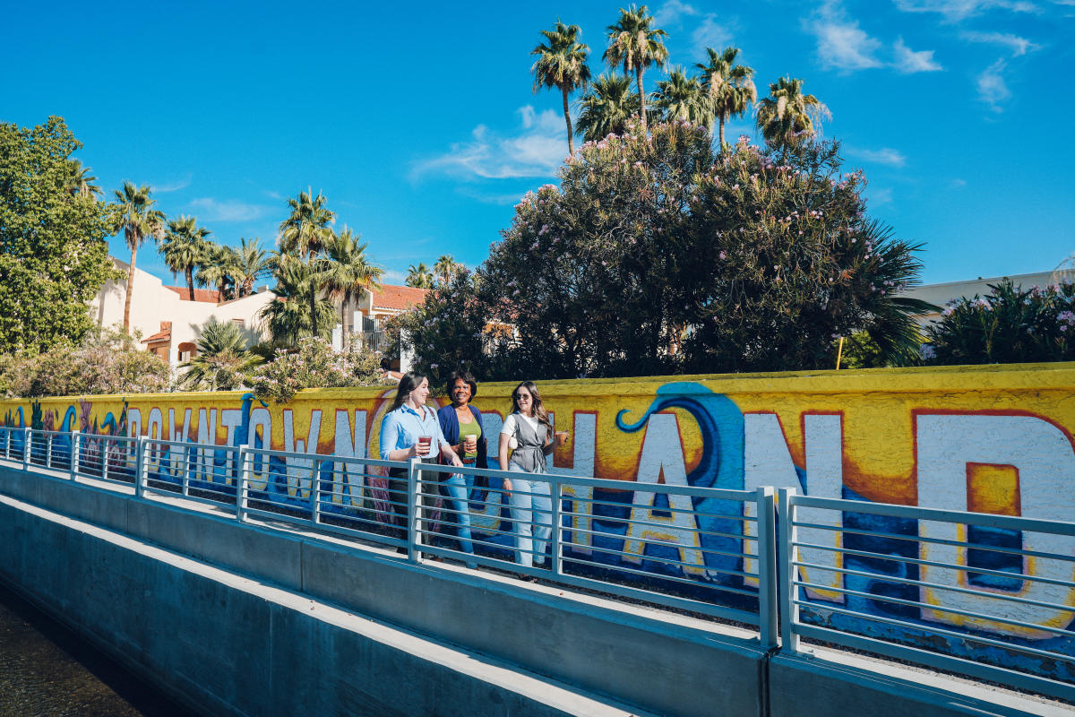 Three ladies walk alongside a bright vibrant mural on a wall that reads "Downtown Chandler" that follows a canal.