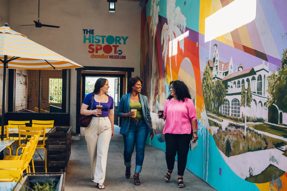 Three ladies walk down a breezeway with a vibrant colorful mural on the wall to the right depicting historic buildings from Downtown Chandler.