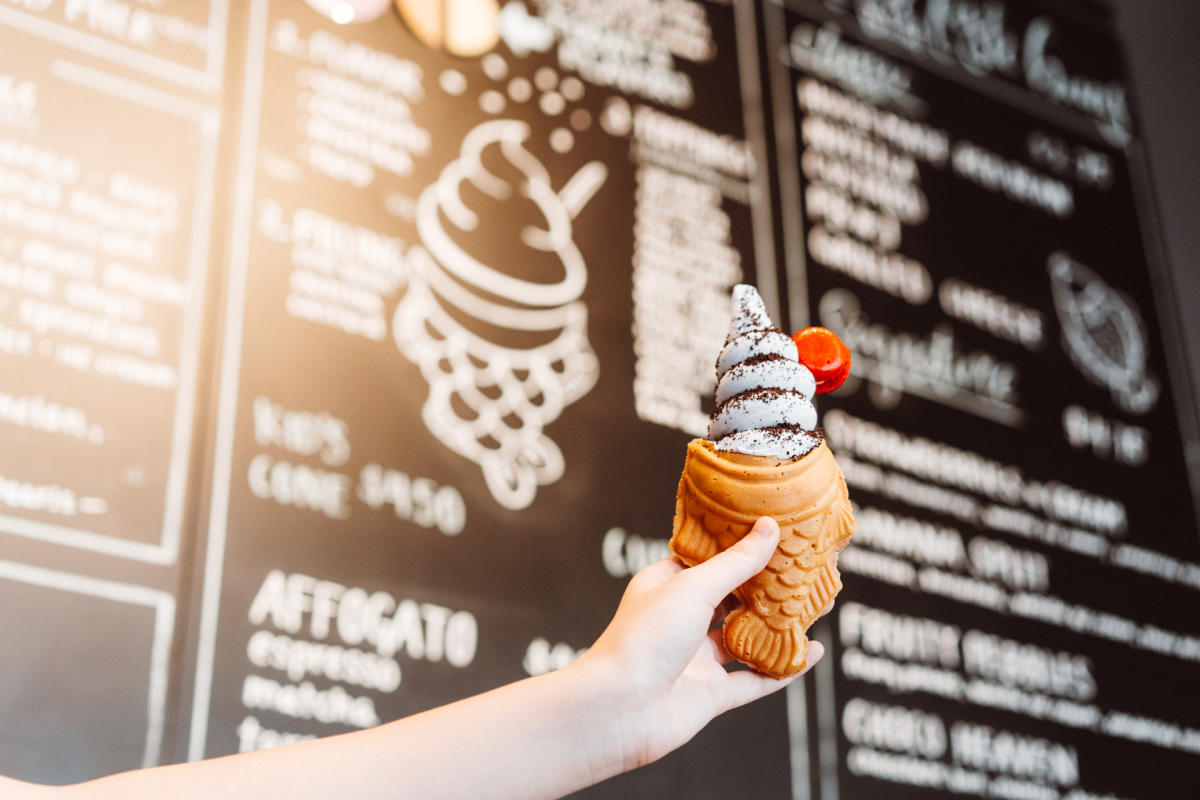 Black menu board with white text blurred in the background with hand in focus holding a fish shaped ice cream cone with swirled ice cream topped with cookie dust and a cherry.