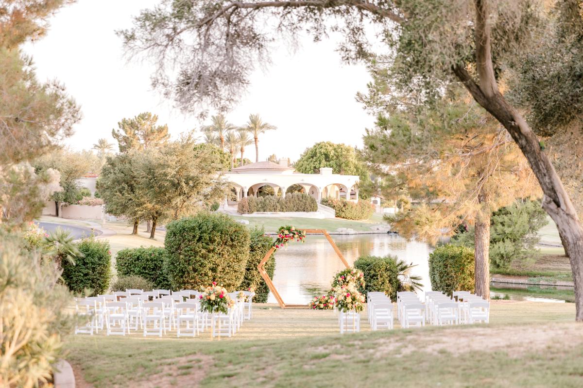 5 rows of white chairs with an isle in the middle sitting on green grass, surrounded by green hills, large bushes at the front, a lake peeking through behind the bushes, and a white building in the background across the lake with large archways. A wooden hexagon arch with floral decor sits in front of the rows of chairs centered with the isle.