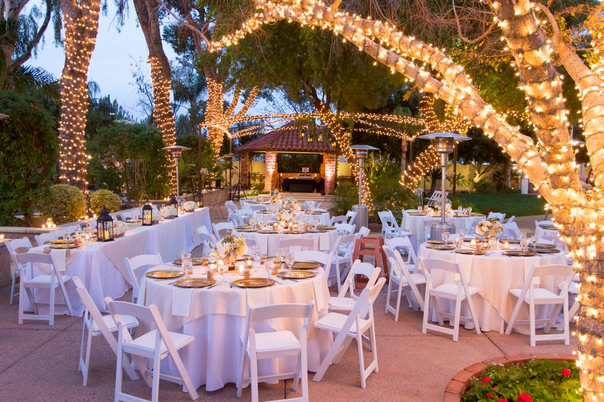 Elegant outdoor wedding setup with lit palm trees, white tables adorned with floral centerpieces, and a cozy ambiance.