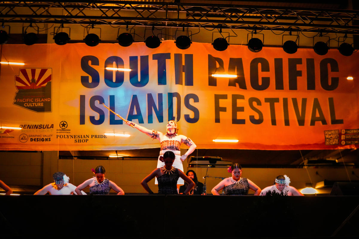 Performers dressed in South Pacific Island cultural attire dance on a stage at night with a banner lit up behind them that reads "SOUTH PACIFIC ISLANDS FESTIVAL".