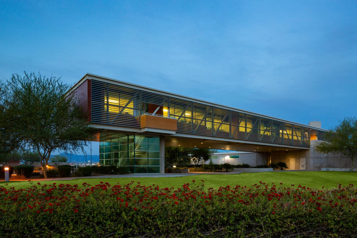 The Chandler Gilbert Community College Williams Hall at dusk, featuring a modern, glass and brick building with a breezeway in the background and a grass and garden in the foreground.