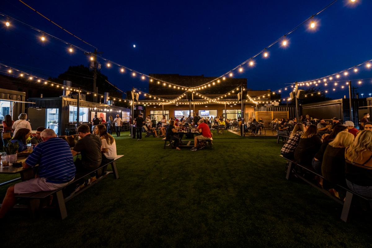Image is of Covington Yard at night with twinkle lights above, people sitting at table and food trucks (operating out of containers) is lining the area.