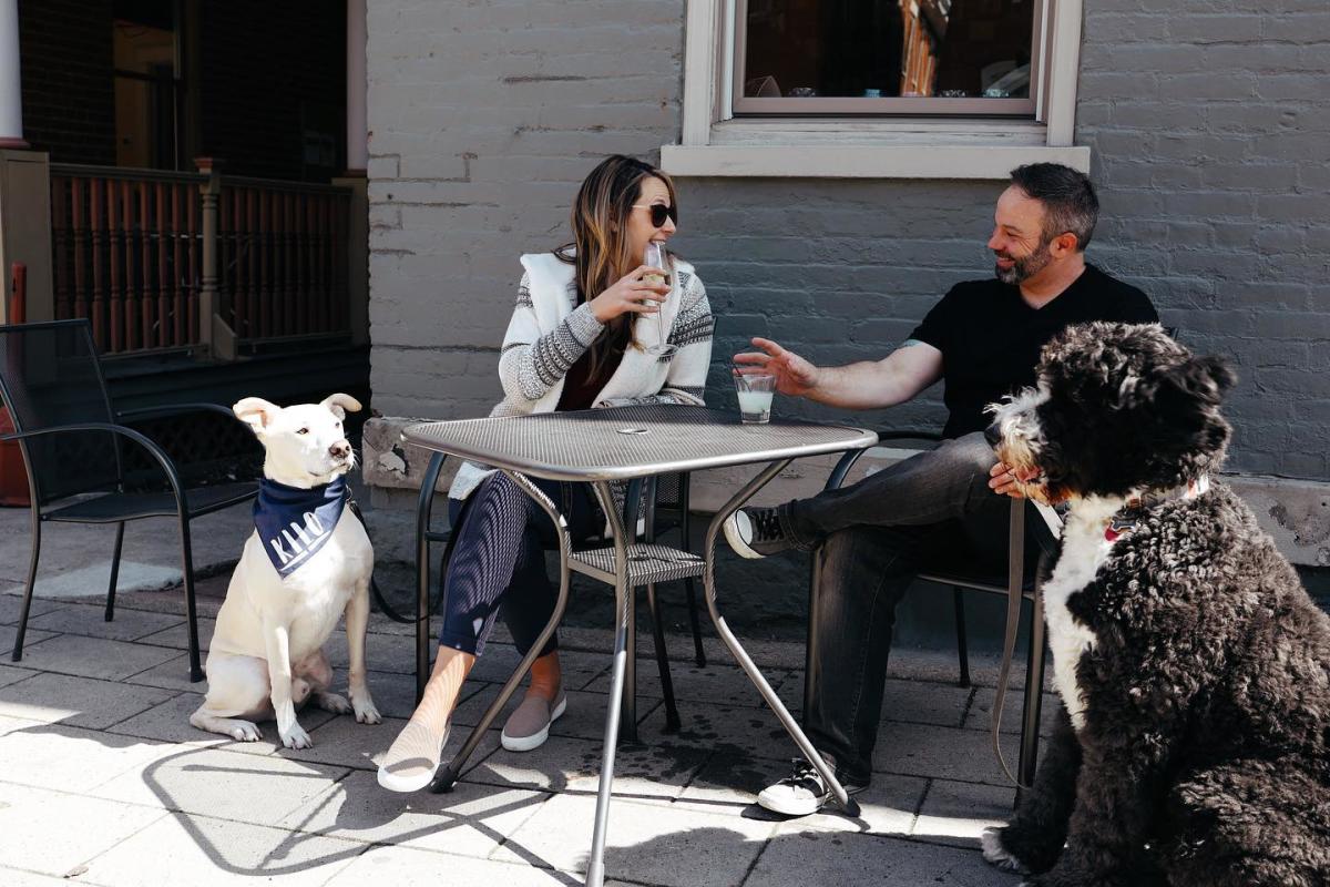 Image is of a couple sitting outside of Craft and Vines with their dogs, enjoying a beer and a glass of wine.