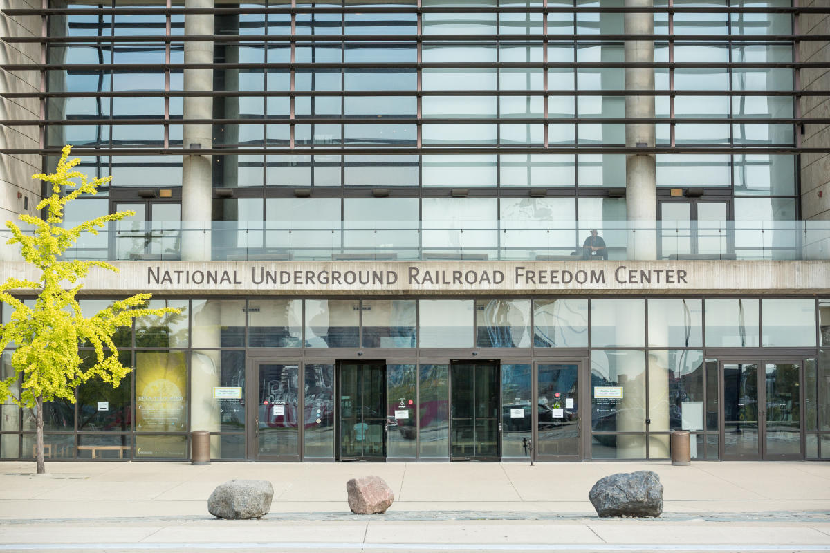 The glass walled front entrance of the National Underground Railroad Freedom Center with a tree, a man, and large stones.