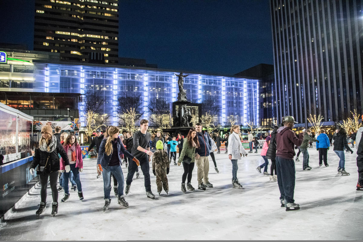 Image is of adults and children, ice skating at Fountain Square in the evening  with trees covered in holiday lights in the background.
