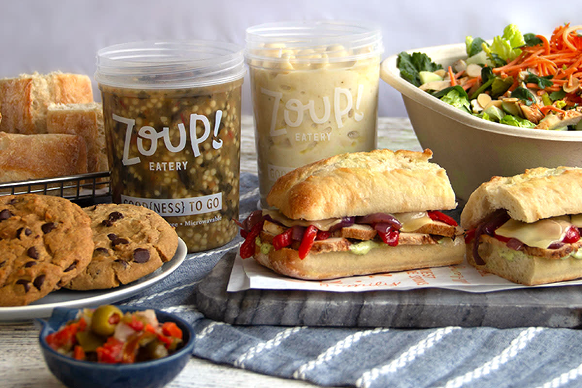 Image is of a display of food containing chocolate chip cookies, salad, bread, sandwiches and clear plastic containers that say Zoup on them with soup inside.