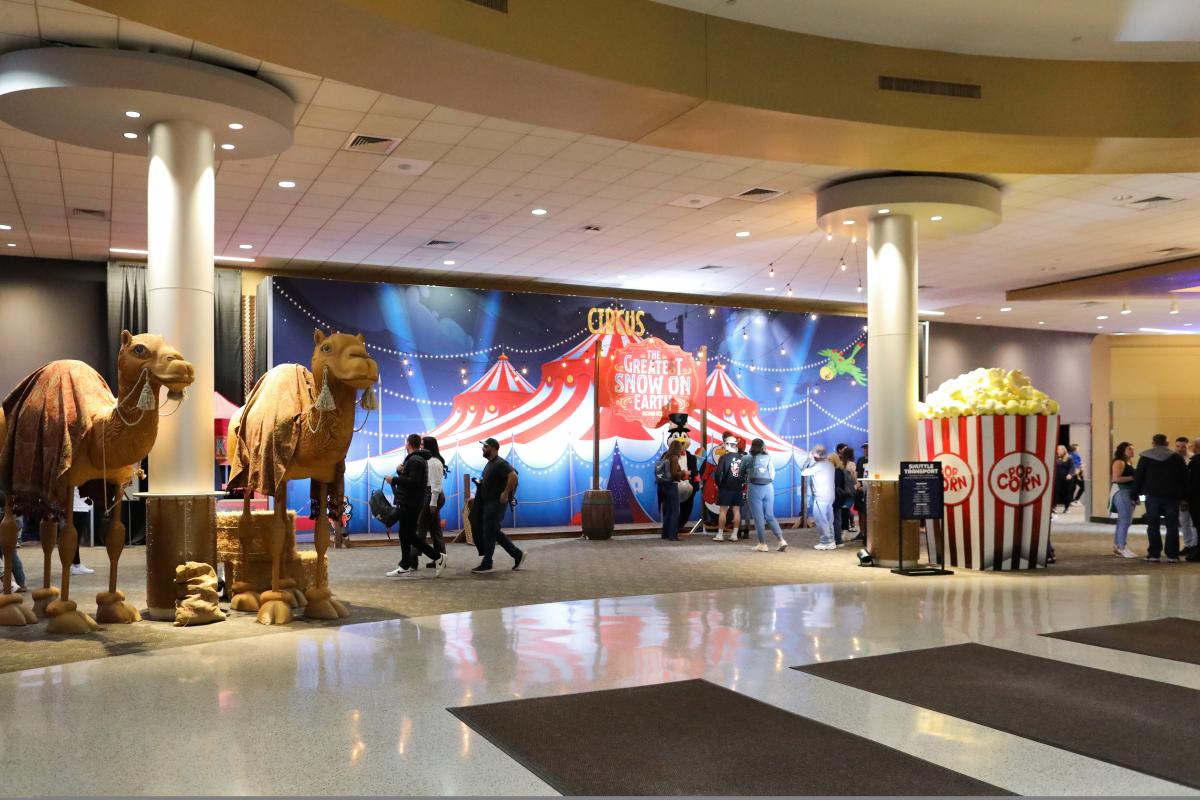 Two statues of camels on the left, a giant popcorn box on the right with people walking past a sign that reads "The Greatest Snow on Earth" over a circus tent painting