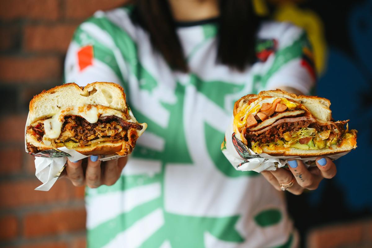A woman in a green and white shirt holding two torta sandwiches out to the camera