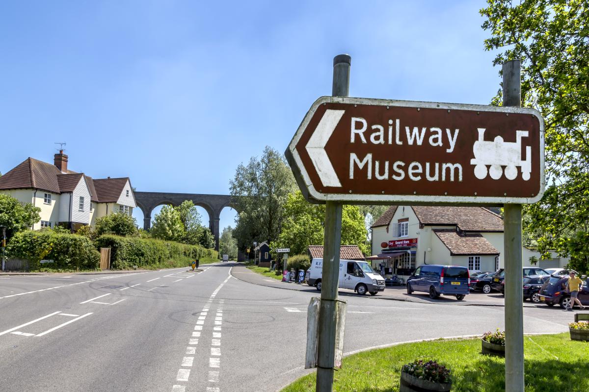A brown roadsign points towards a Railway Museum. Behind it is a main road, some picturesque houses and a large brick viaduct.