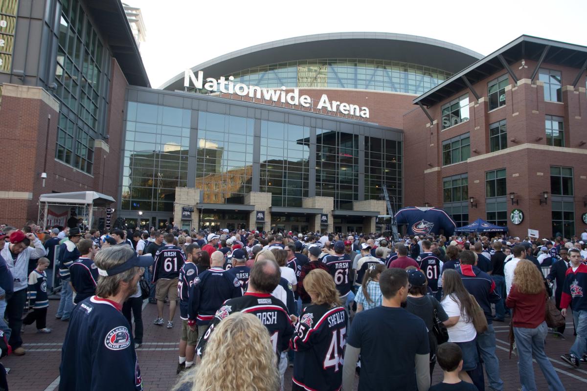 Crowd wearing Columbus Blue Jackets jerseys gathers outside Nationwide Arena with glass windows and brick facade.