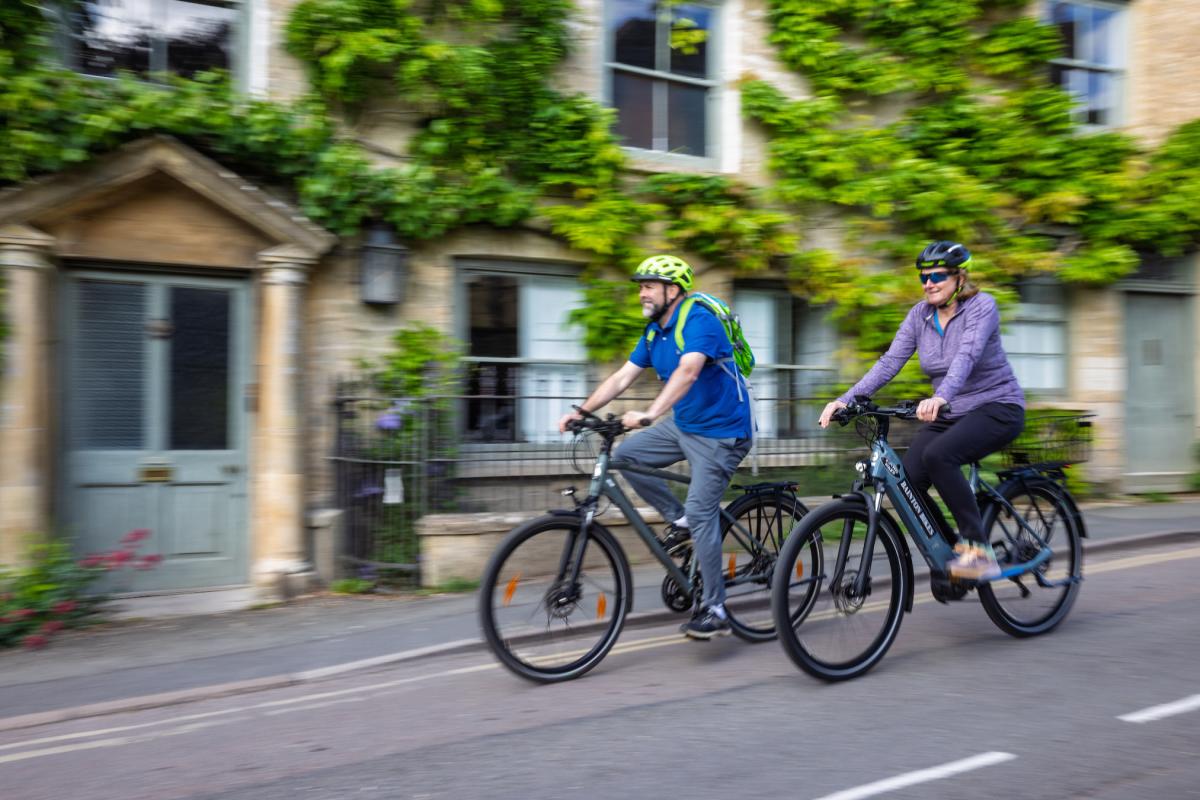 Two cyclists riding bikes through picturesque Charlbury, passing stone cottages covered with greenery