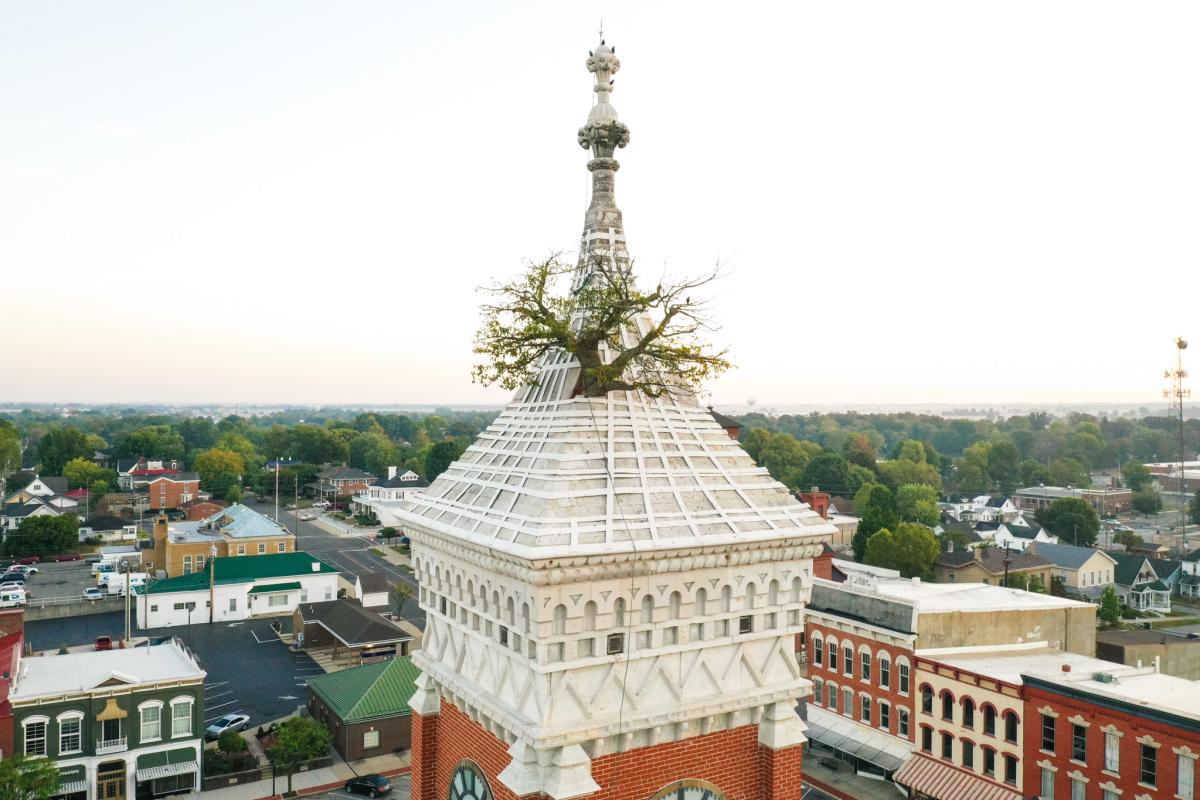 Aerial view of Greensburg, Indiana’s historic Decatur County Courthouse, featuring the famous Tower Tree growing from the courthouse roof, with the downtown area and surrounding landscape visible in the background.