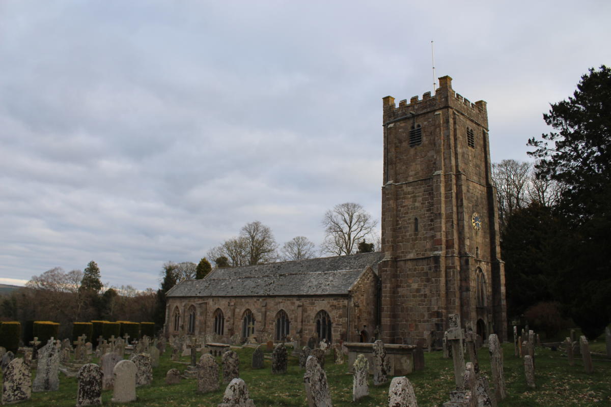 Stone church with a tall tower and clock set amid an old cemetery. Overcast sky and leafless trees create a somber, historical atmosphere.