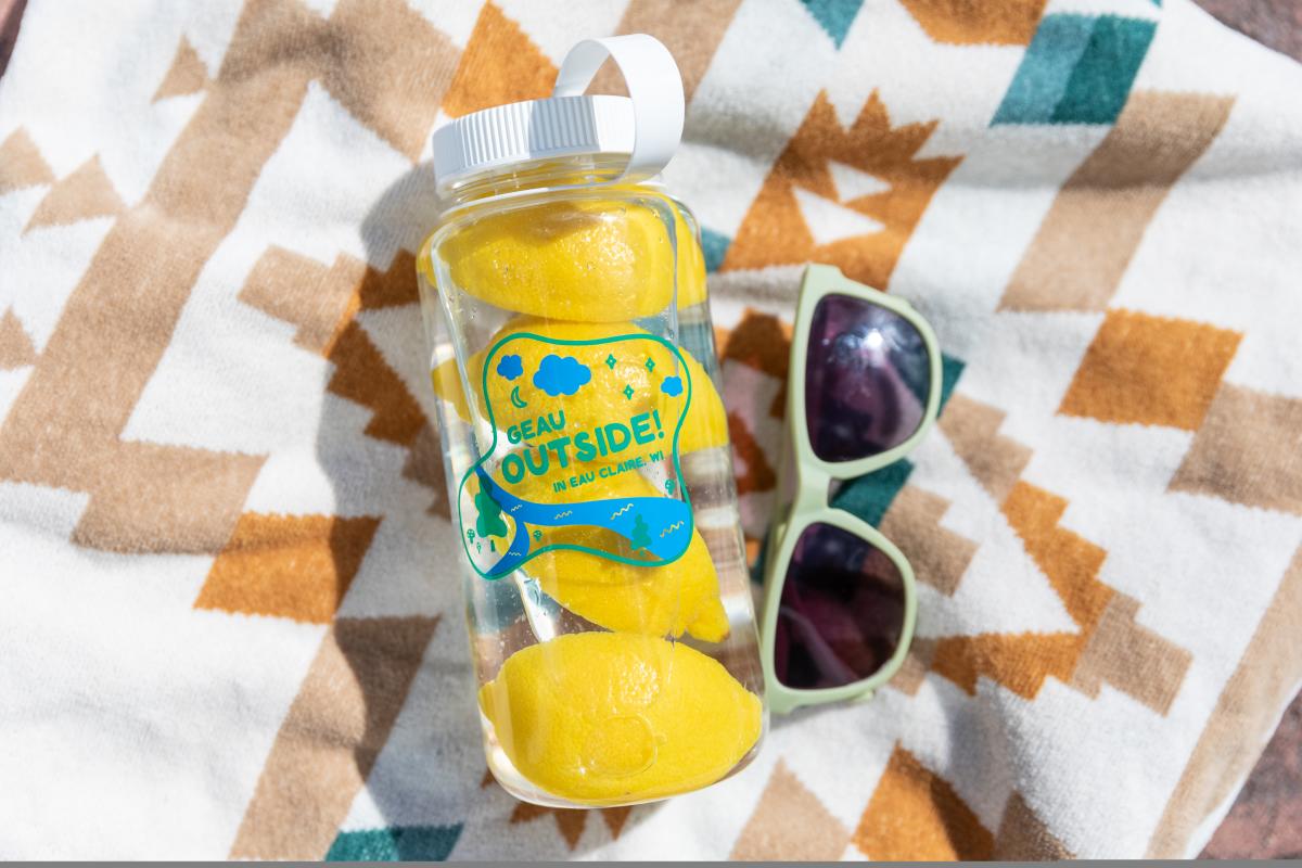 A photo of a clear water bottle filled with lemons and water next to some sunglasses on a blanket