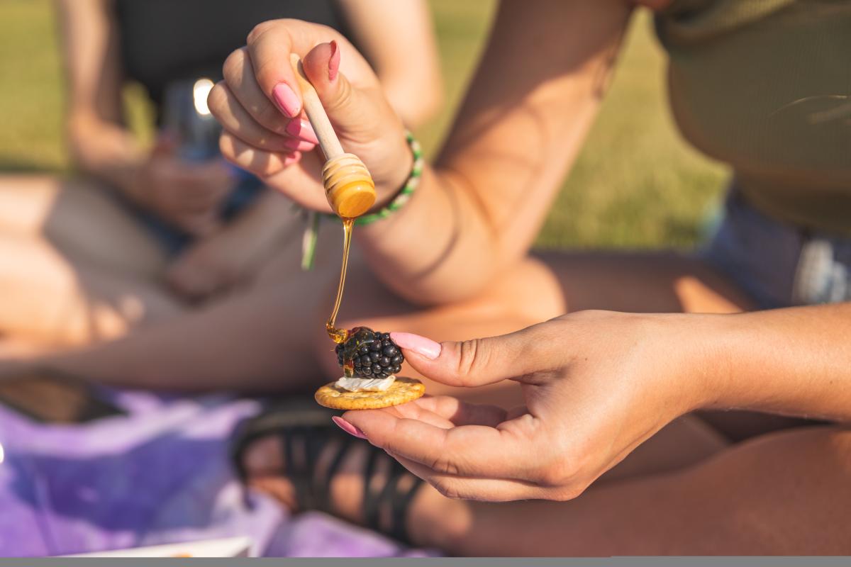 A girl putting honey on a cracker with cheese and a blackberry