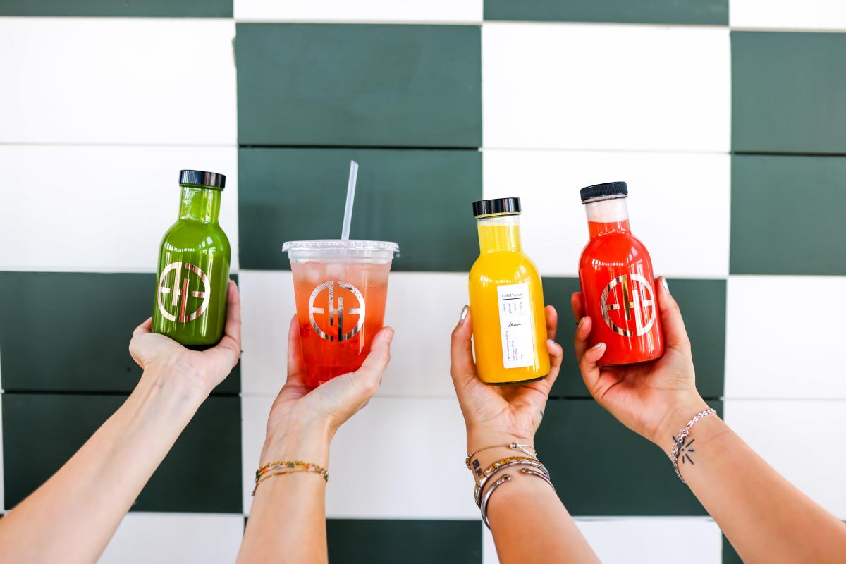 Four hands holding up fresh green, yellow, and red juice in front of a checkered print wall at Harlow Juice Bar