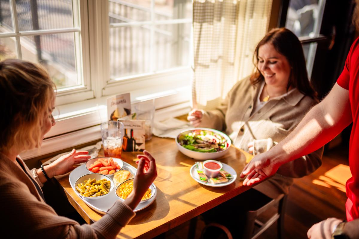 Two women enjoying Southern food at Back Home restaurant as a server delivers a dish, showcasing local flavors in a cozy, sunlit setting.