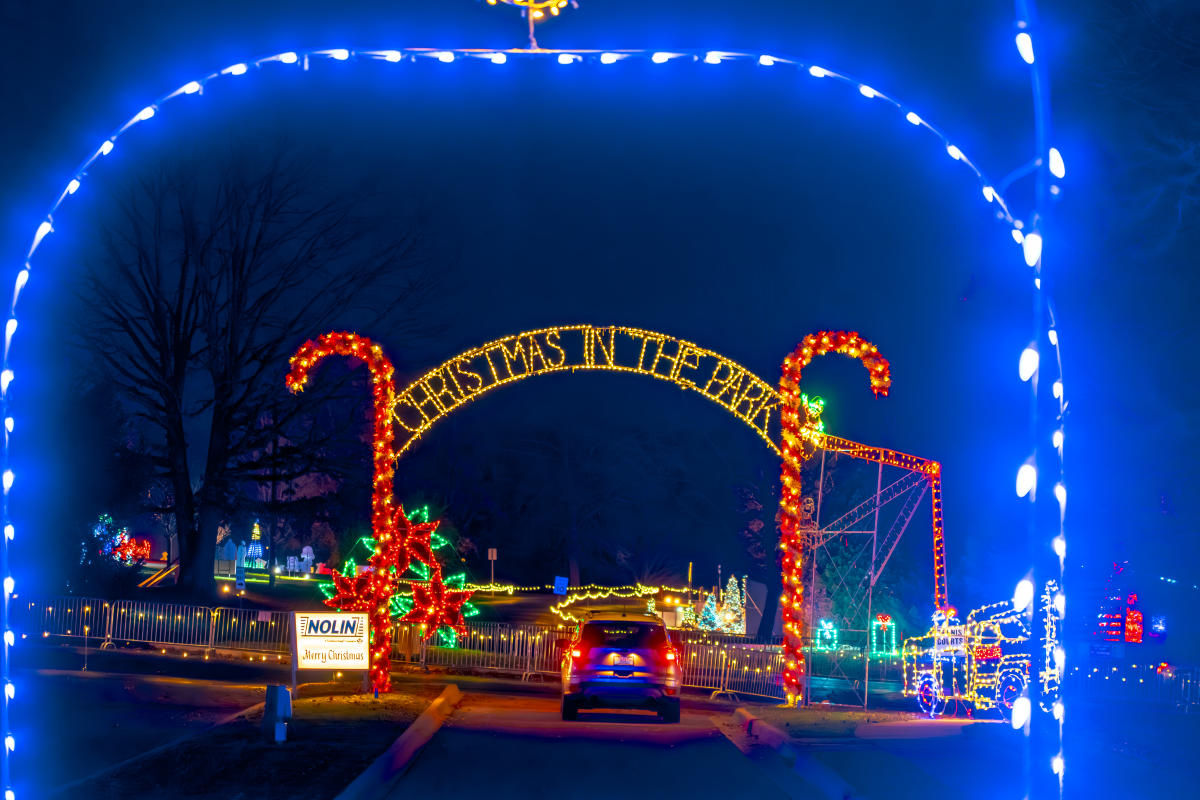 Entrance to a festive holiday light display, featuring an archway decorated with glowing candy canes and colorful Christmas lights, surrounded by illuminated decorations and trees.