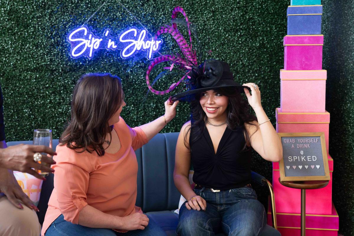 A woman is reaching out and touching a Kentucky Derby hat on top of another woman's head while sitting down on a couch in That Cute Little Shop in Downtown Elizabethtown