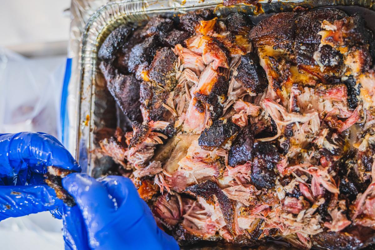 A close-up of shredded, smoky pulled pork in a tray, with someone wearing blue gloves preparing or serving the meat.