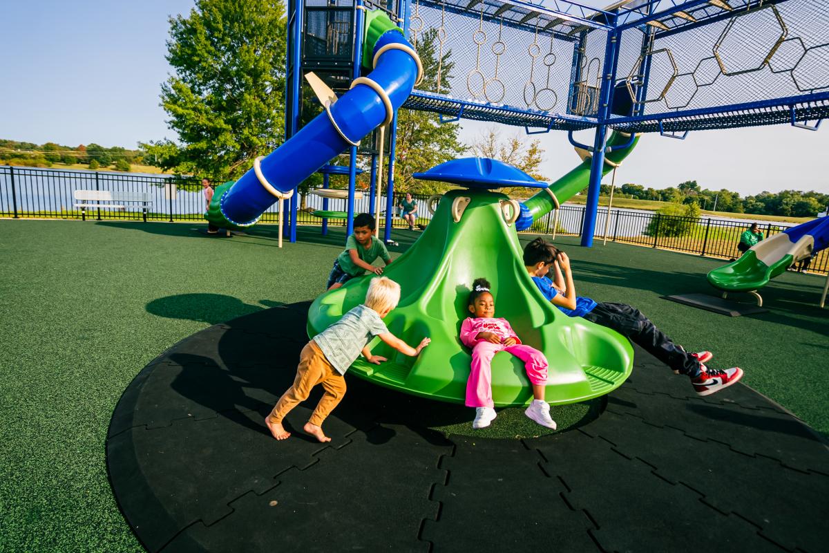 Children playing on a green spinning structure at a playground with a large blue slide in the background.