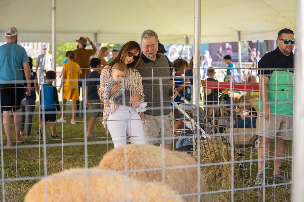 A family with a young child observing animals in a petting zoo under a tent at an outdoor event, with other people and children in the background.