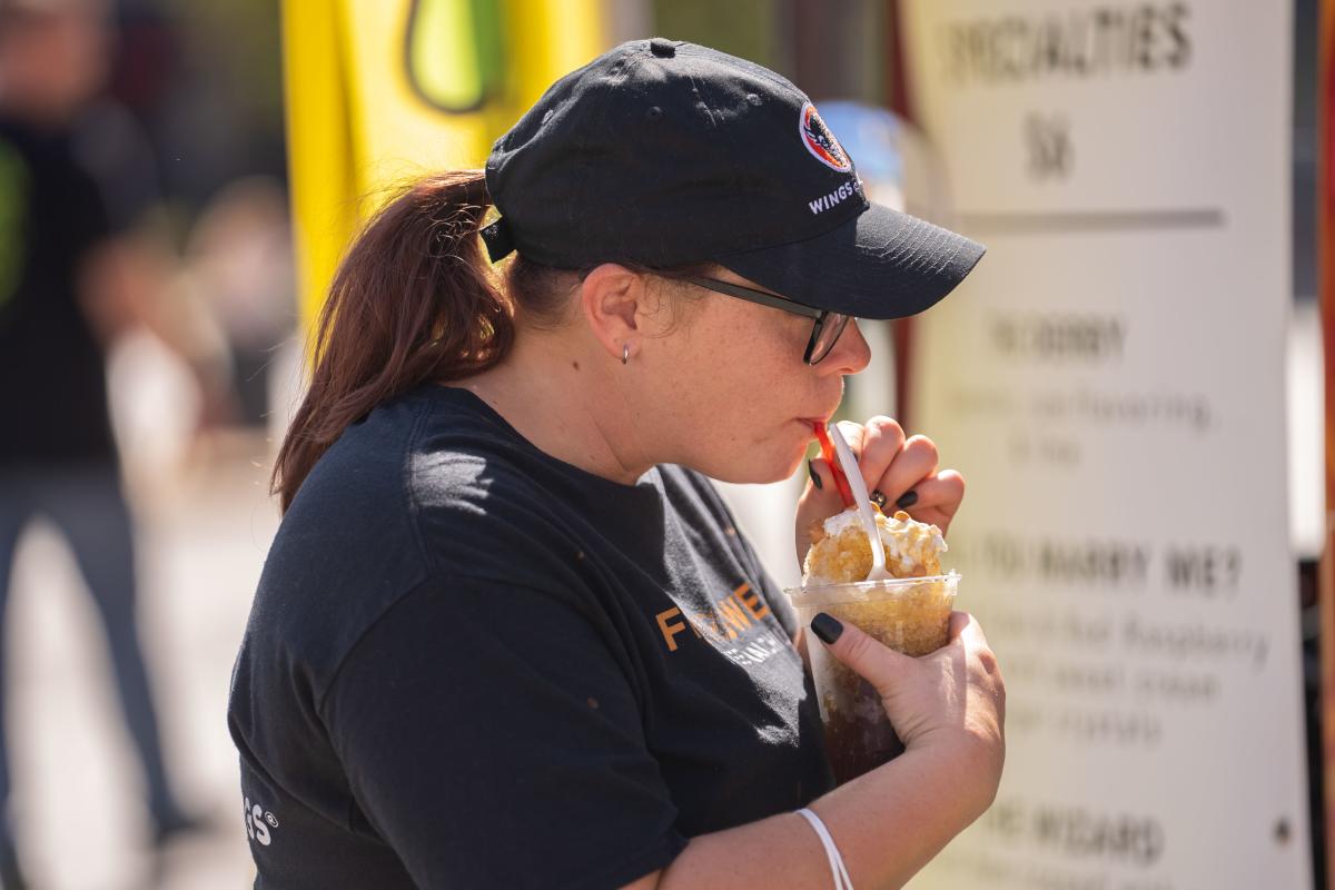 A woman wearing a black cap and shirt enjoying a dessert from a small cup with a spoon while standing near a food vendor at an outdoor event.