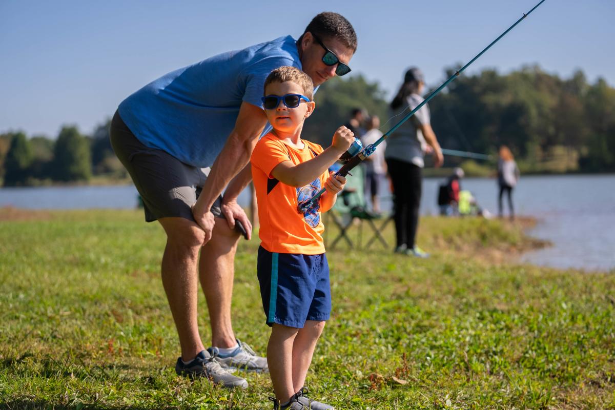 A father and son wearing sunglasses and fishing together by a lake on a sunny day, with other people fishing in the background.