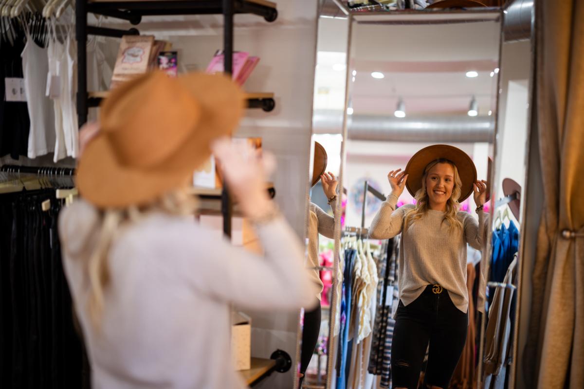 Person smiling while trying on a stylish brown hat in front of a mirror at a clothing store.