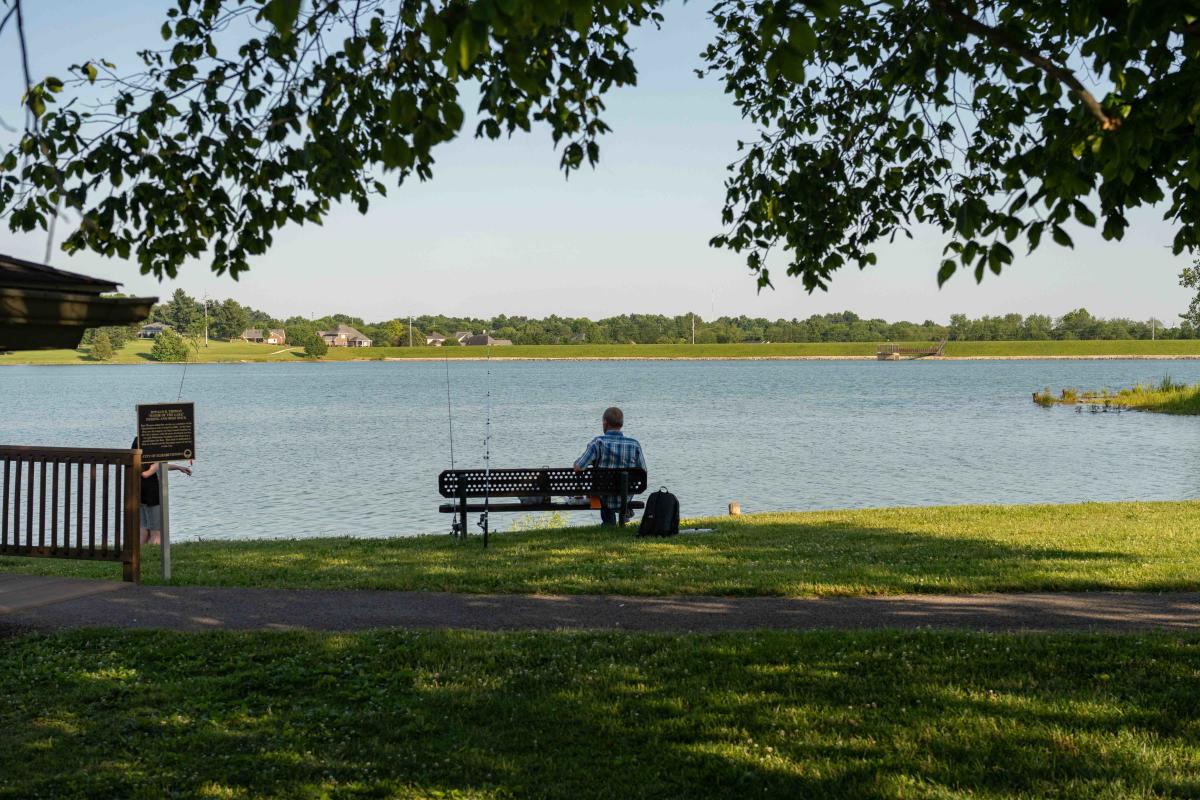 A man fishing at Freeman Lake in Elizabethtown, KY, sitting on a bench by the calm water under a clear sky.