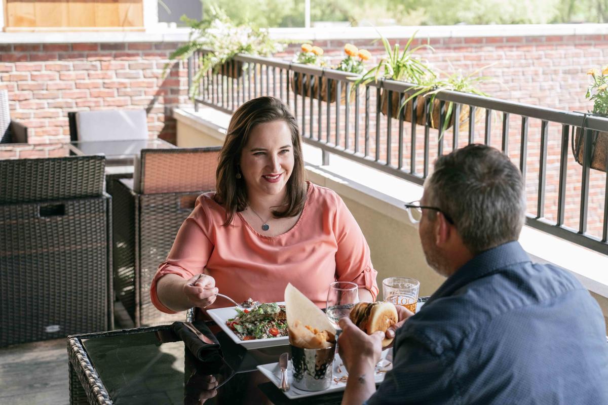 Woman smiling at the man sitting across from her on the patio of J.R. Neighbors. She is also eating her salad with salmon on top, and he is eating a burger with a side of tater tots