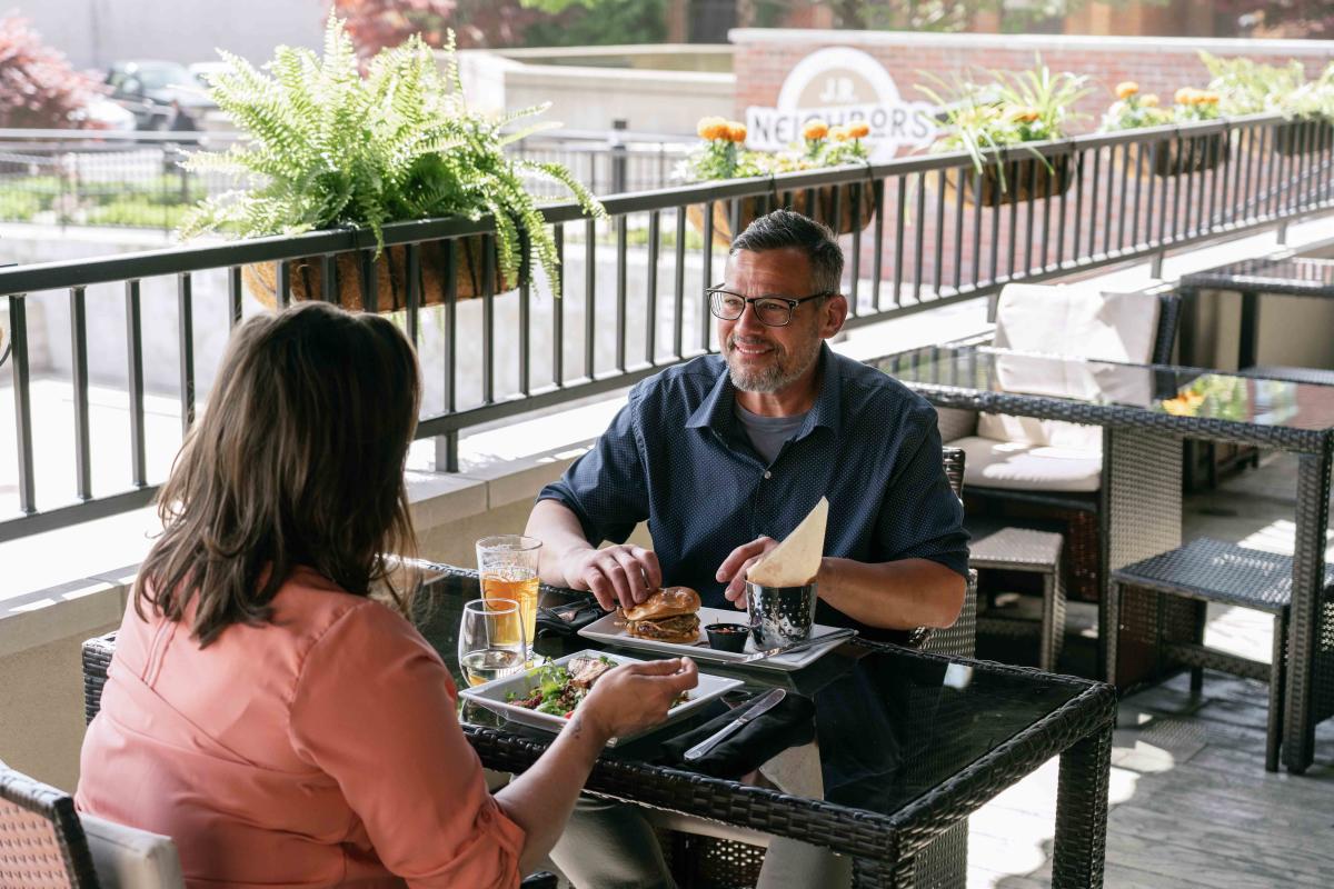 A couple enjoys a meal together on a sunny outdoor patio, surrounded by plants and a relaxed atmosphere.