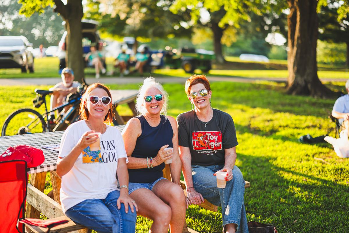 Three ladies are smiling while sitting down on a picnic table in Freeman Lake listening to the band at the Lawn Party in Elizabethtown, Kentucky