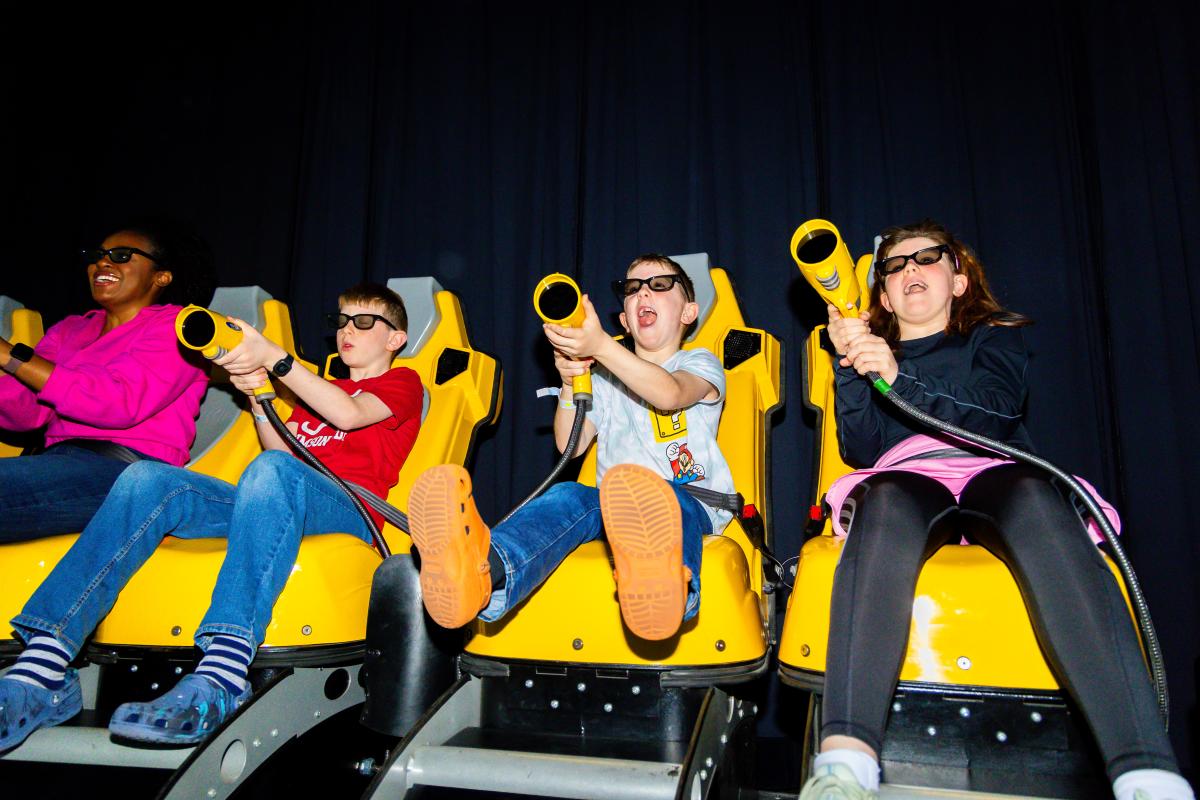 Kids and adults wearing 3D glasses, sitting in yellow motion seats, and using interactive blasters in a virtual gaming experience.