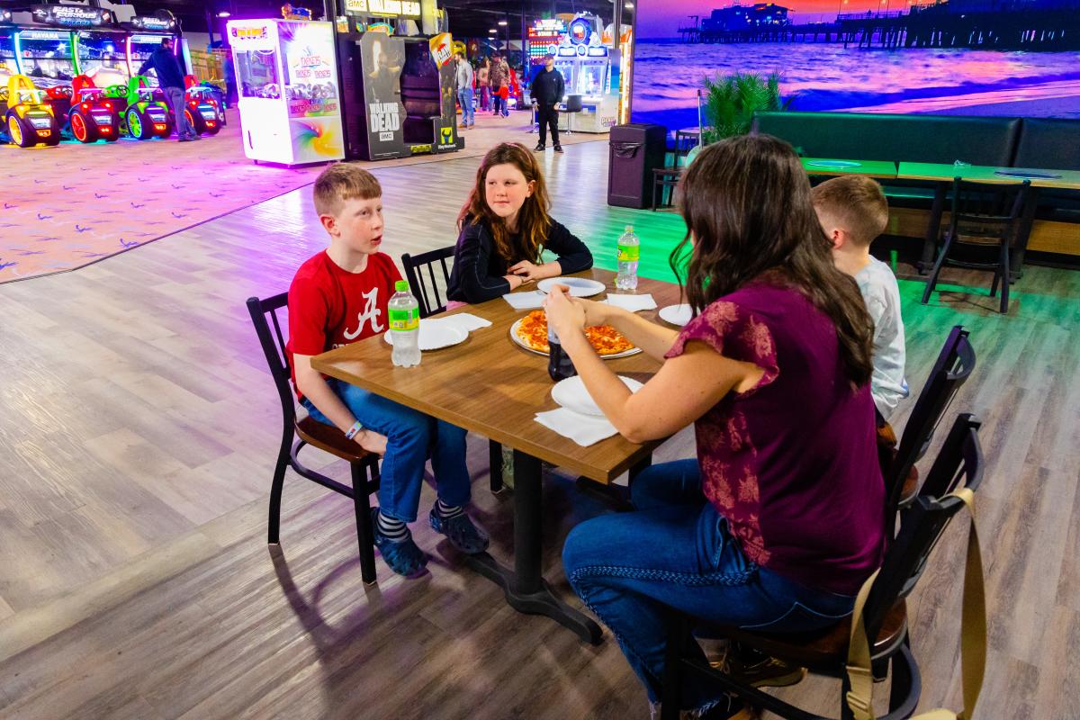 Family sitting at a table enjoying pizza and drinks inside an arcade with colorful games in the background.