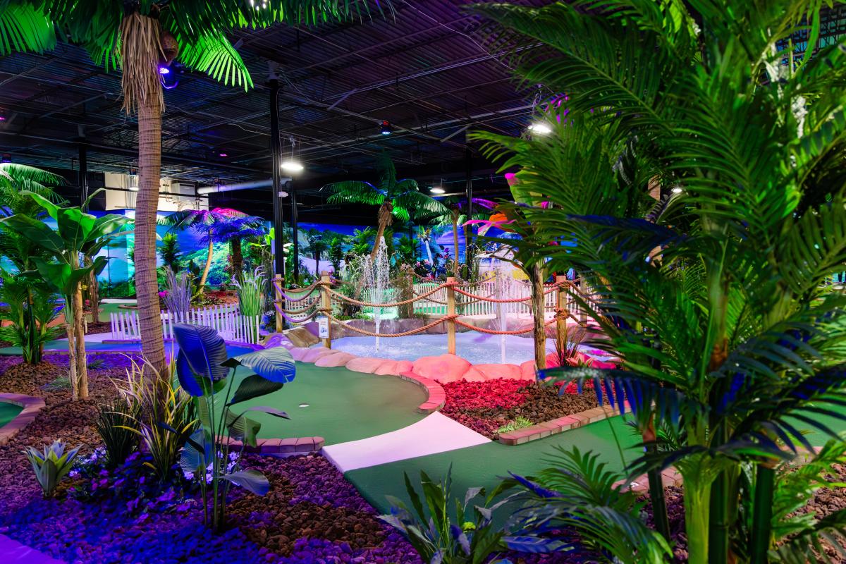 Indoor mini-golf course surrounded by lush tropical plants, vibrant lighting, and decorative features.