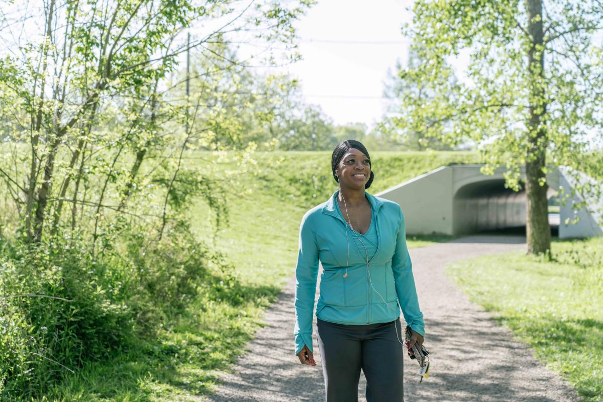 A woman walks along a tree-lined path on a sunny day, smiling and enjoying the outdoors.