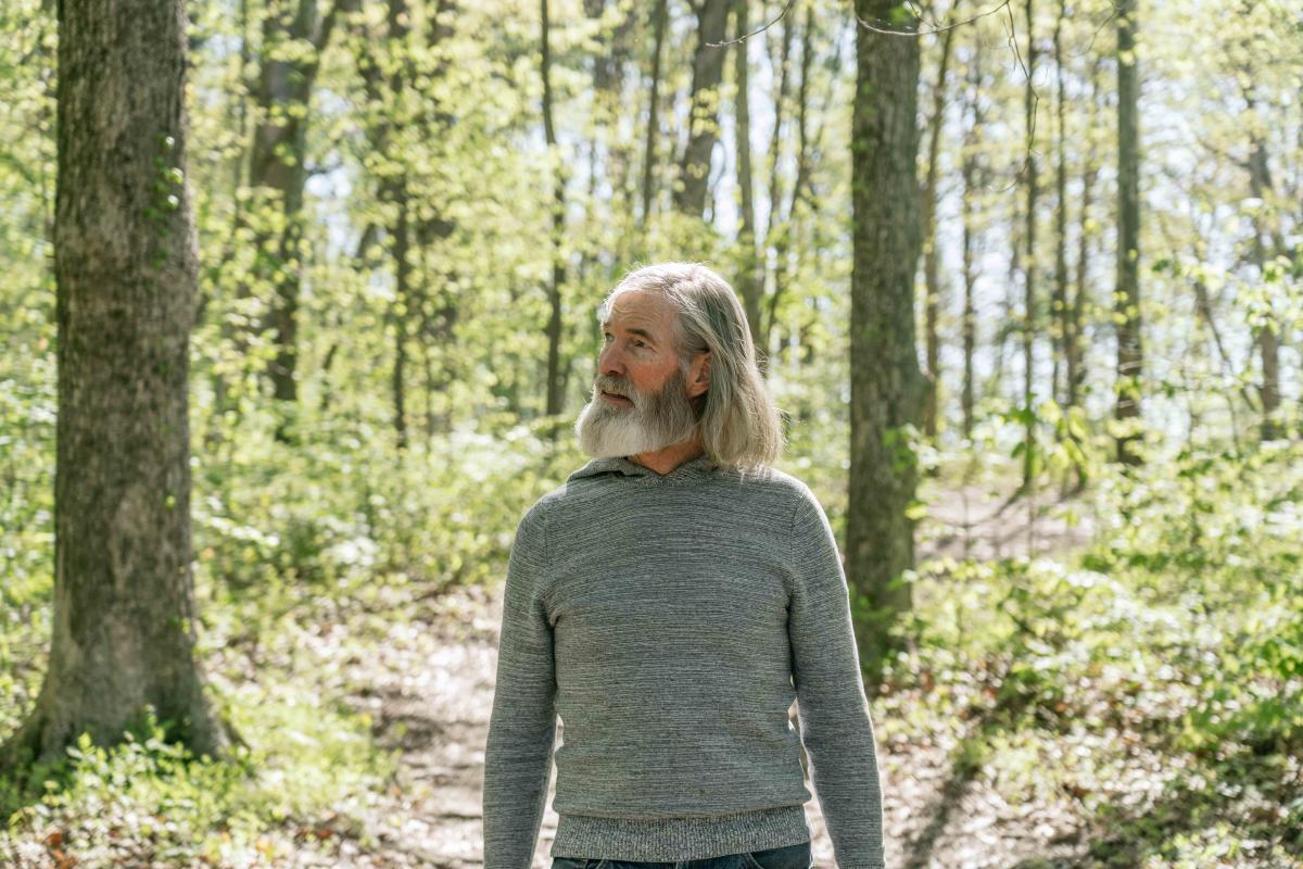 A man with long gray hair and a beard walking through a sunlit forest, wearing a gray sweater.