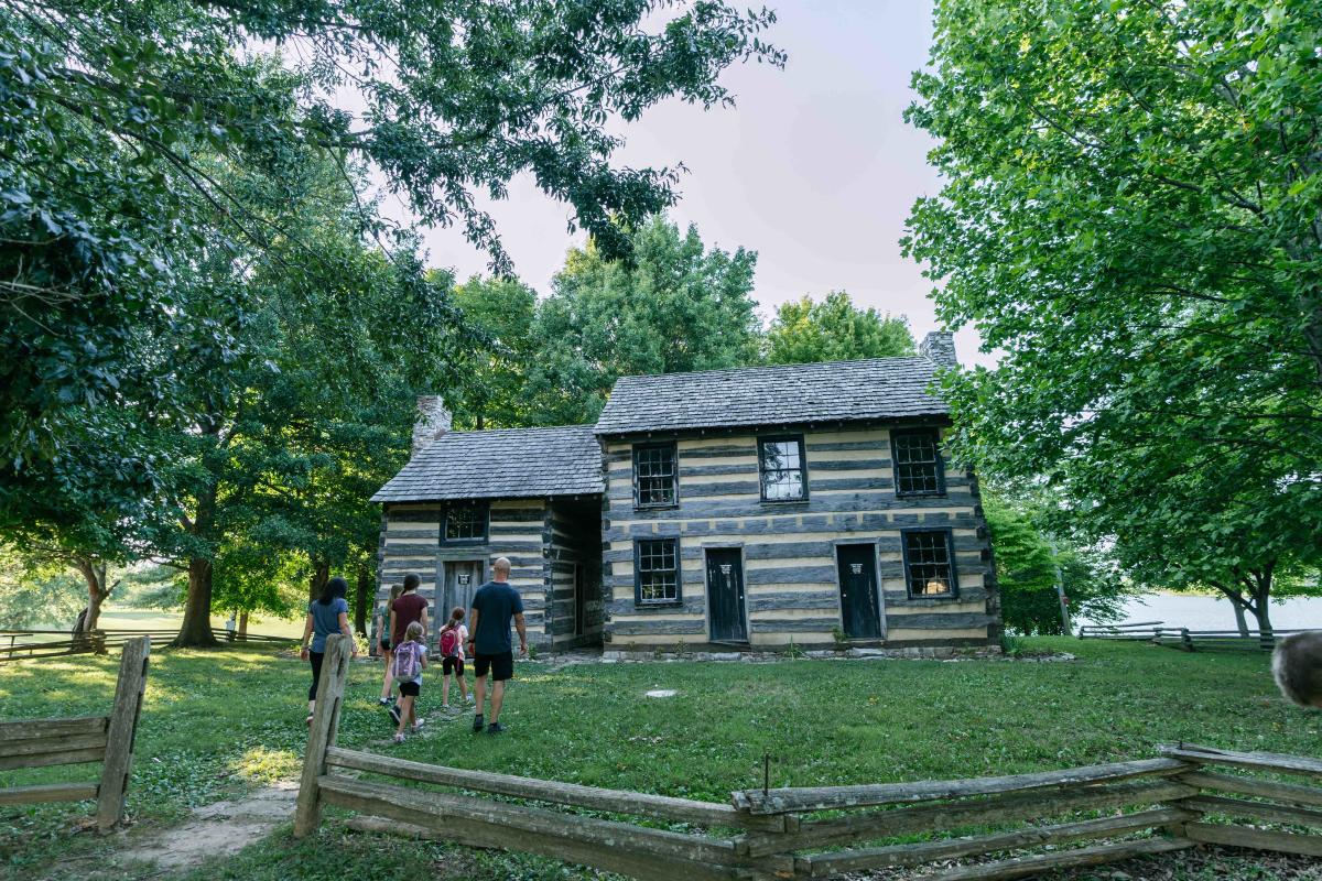 A historic two-story log cabin surrounded by lush green trees, with a small group of visitors, including adults and children, walking towards it on a grassy yard, enclosed by a wooden fence.