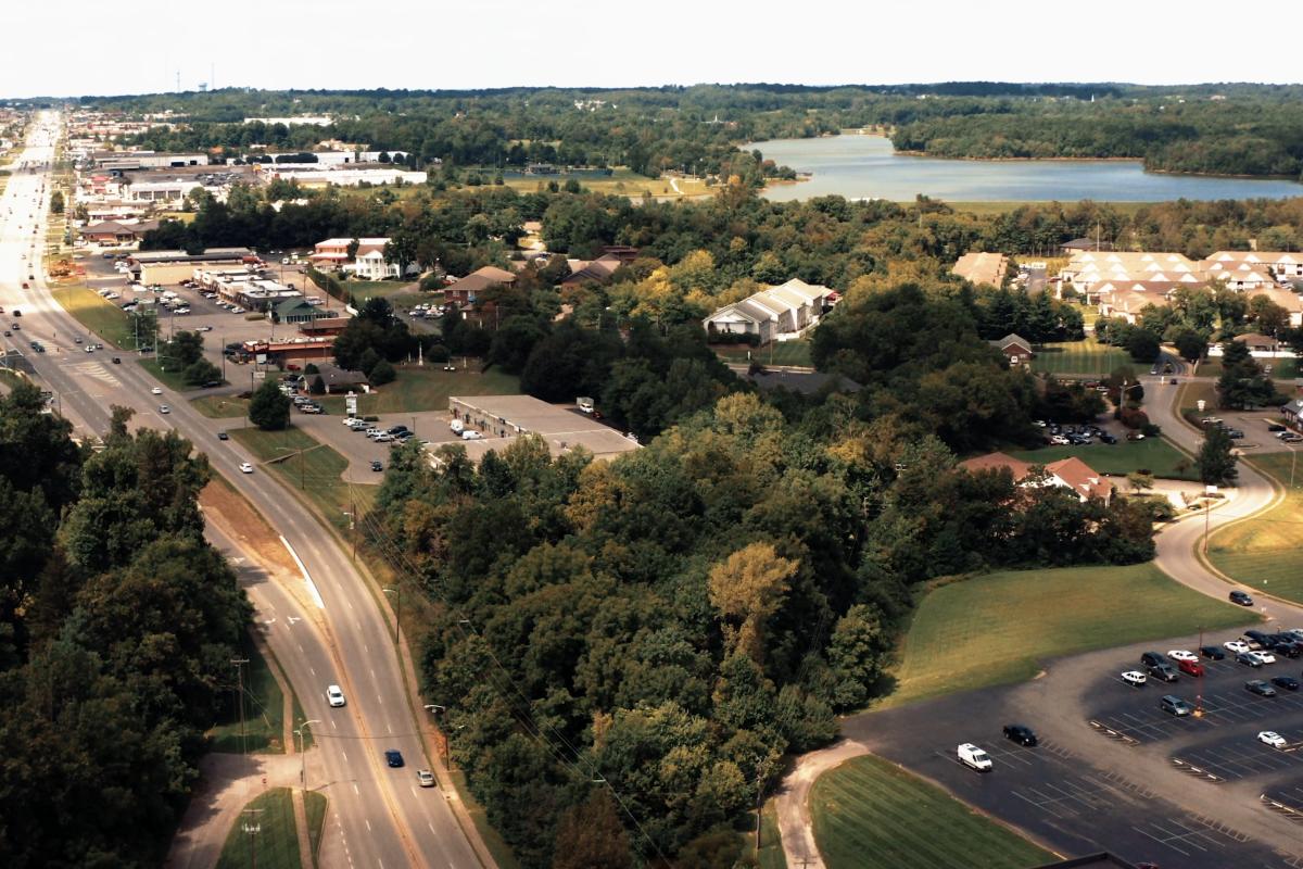 Aerial view of roads, buildings, trees, and a lake in the distance, capturing a mix of natural and developed areas.