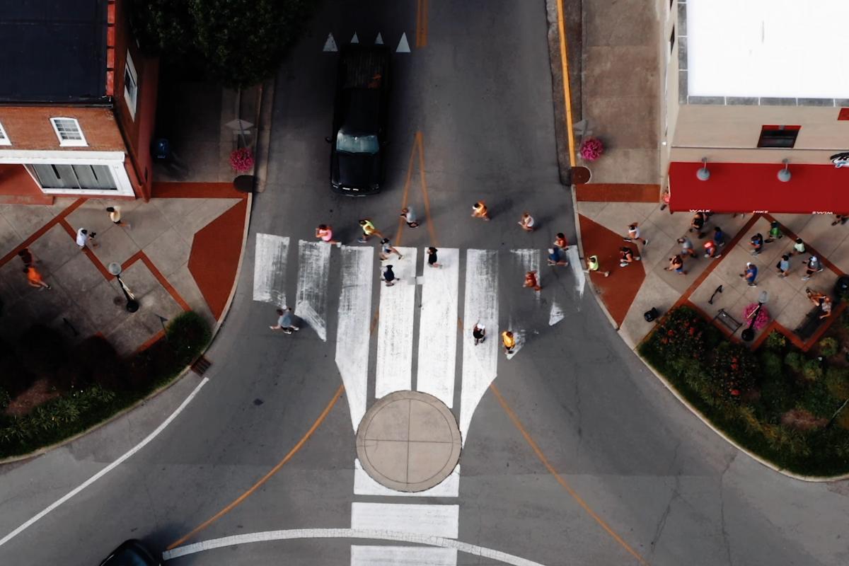 Aerial view of a crosswalk at a busy intersection with people walking across and cars waiting, surrounded by buildings and outdoor seating areas.