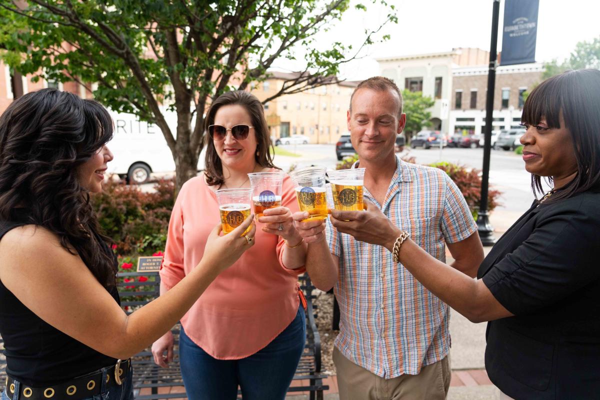 Three women and one man are holding up their Hub Cups in a cheers while on the sidewalk in Downtown Elizabethtown
